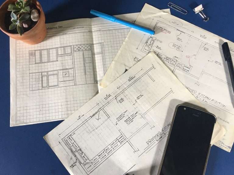 Blueprints for home addition and kitchen remodeling with a plant, pen, and smartphone on a blue background, illustrating the planning phase of construction services.