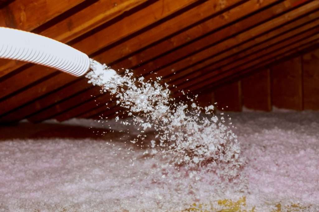 Technician spraying blown fiberglass insulation in an attic, enhancing energy efficiency for homes with black shingles.