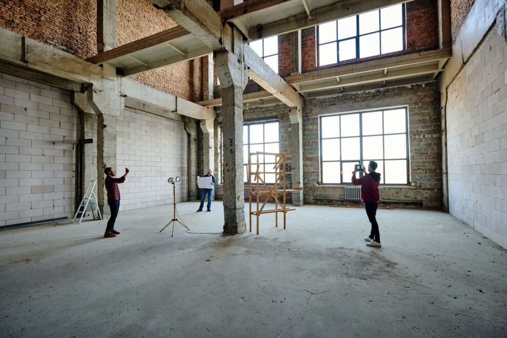 Two young contractors photographing an unfinished structure inside a spacious building, showcasing the construction process and potential for home additions.