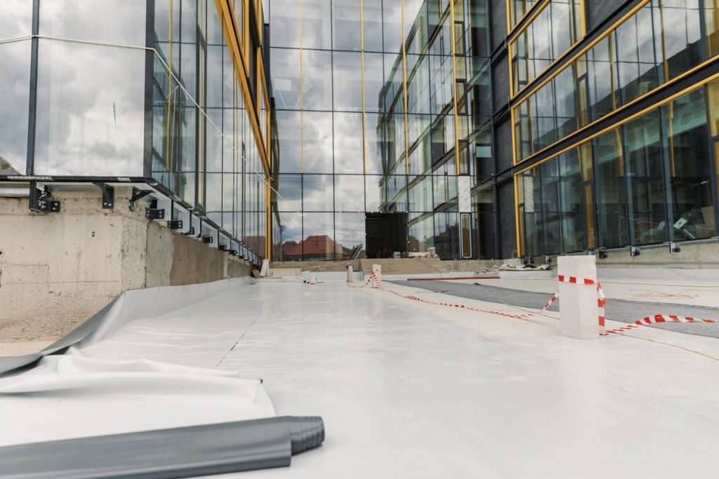 Construction site featuring a flat roof installation with a white membrane, surrounded by modern glass building facades and safety barriers, illustrating commercial roofing solutions.
