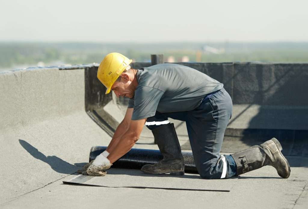 Worker installing single-ply membrane roofing on a commercial building, wearing safety gear including a hard hat and gloves, demonstrating roofing techniques relevant to commercial roofing solutions.