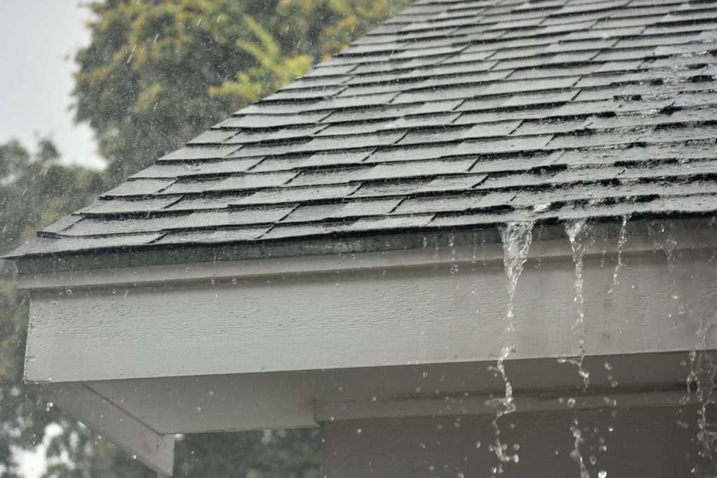 Roof with asphalt shingles during heavy rain, showcasing water runoff along the eaves, highlighting potential signs of water damage related to hail impact.