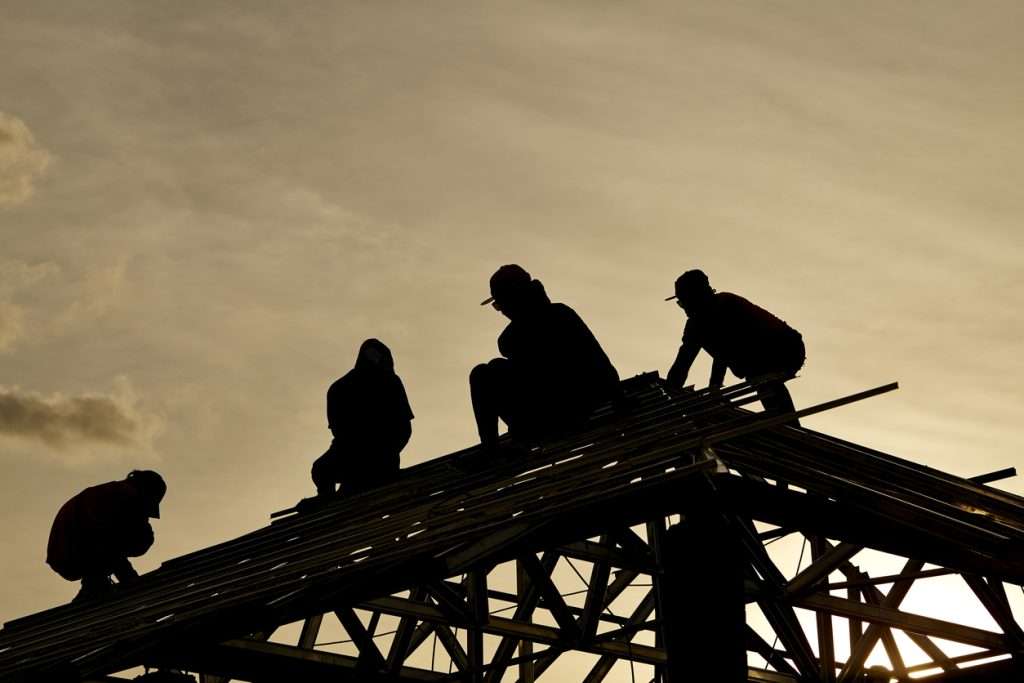 Silhouetted construction workers installing roofing materials on a commercial building at sunset, highlighting the labor involved in roofing projects.