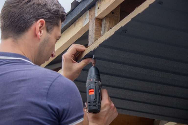 Man repairing a roof, tightening a self-tapping screw with a power drill, emphasizing roofing maintenance and construction services.