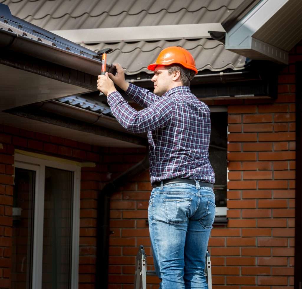 Man in an orange hard hat installing gutters on a residential roof, showcasing expert construction work for water damage prevention.