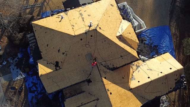 Aerial view of a residential roof under construction, showcasing workers installing new roofing materials on a multi-pitched roof, with blue tarps covering the ground for protection.