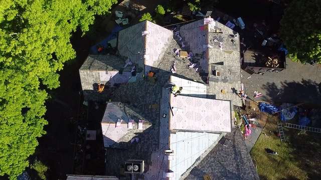 Aerial view of a residential roofing project in progress, showcasing workers installing new shingles on a multi-pitched roof surrounded by trees, reflecting Artisan Roofing & Construction's emergency repair services.
