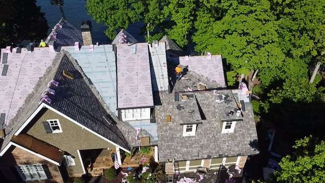 Aerial view of a residential roofing project, showcasing multiple roofs under construction with new shingles and roofing materials, surrounded by greenery and water, highlighting the roofing services offered by Artisan Roofing Construction.