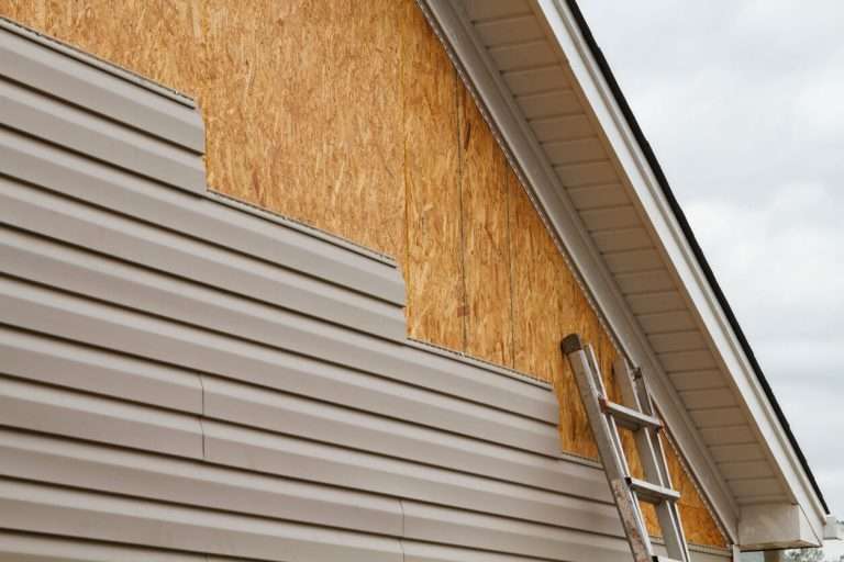 Exterior siding installation in progress, showcasing vinyl siding and wooden sheathing, with a ladder positioned against the house.